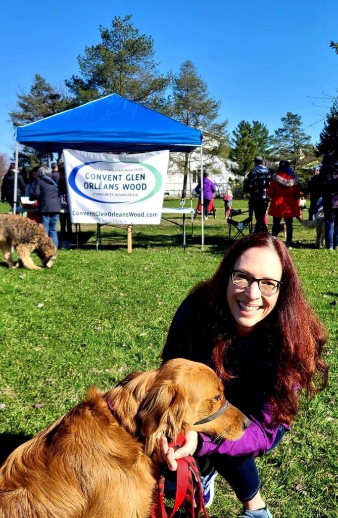 A photo from the 2022 Dog Walkathon.  Laura Dudas attended and is seen with a golden retriever.  In the background, you can see the Convent Glen Orléans Wood banner and more than 10 people, along with some dogs at the park.