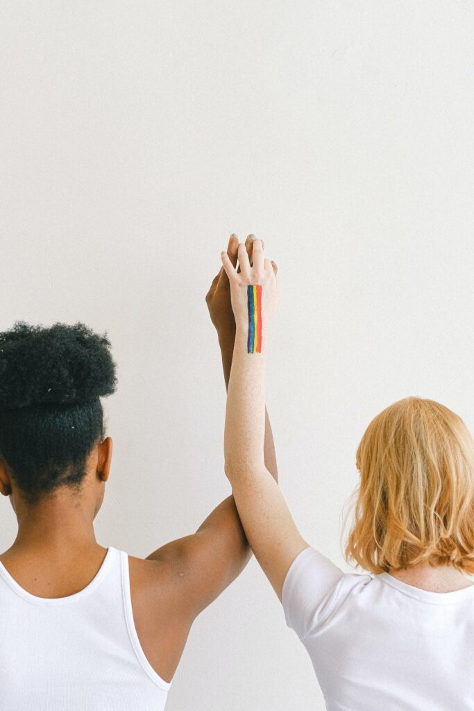 two women holding hands with arms raised. one woman has a rainbow painted on her skin.