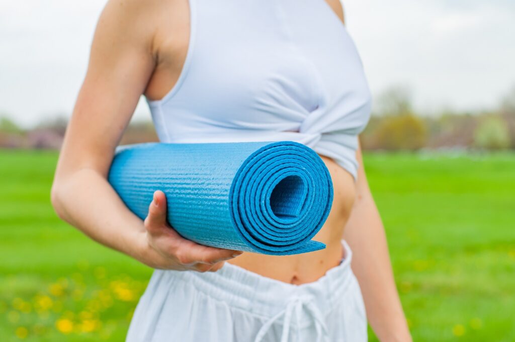 A person wearing white pants and a white tank top who has a rolled up blue yoga mat rolled up and in their arm. They are standing in a park with green grass.