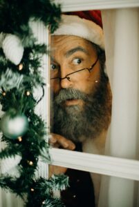 photo of Santa Claus looking out a window of a house that is decorated with garland and christmas lights