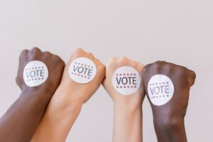 four hands in a fist that are lined up in a row displaying the backs of their hands. There is a sticker on the back of each hand that says vote. Each hand has a different skin tone.