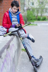 a teenager resting on a ledge with a scooter. The child is looking at a smartphone.