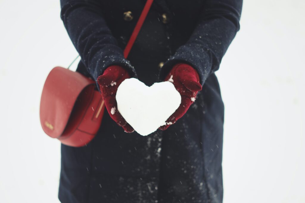 photograph of a person wearing a blue winter coat, red mittens and a red purse who is holding a snowball that is shaped like a heart.