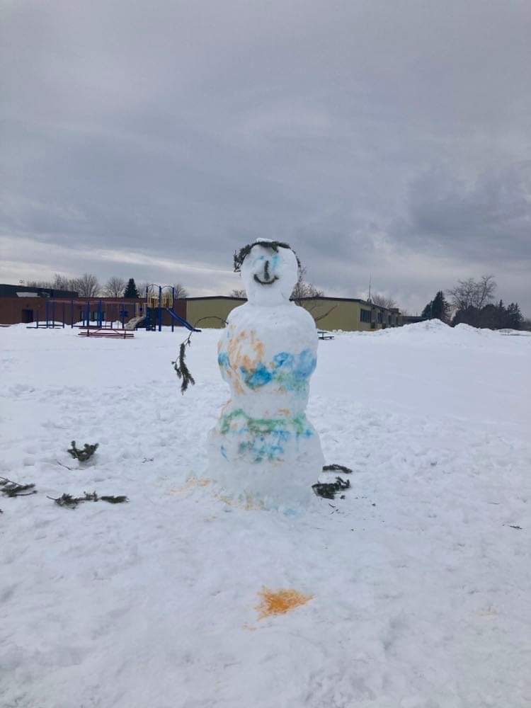 a final completed snowman at the winter carnival. it is made of three snow boulders, it is painted with food colouring, and has branches to make a face, arms and a crown. / un bonhomme de neige terminé au carnaval d'hiver. il est fait de trois blocs de neige, il est peint avec du colorant alimentaire, et a des branches pour faire un visage, des bras et une couronne.