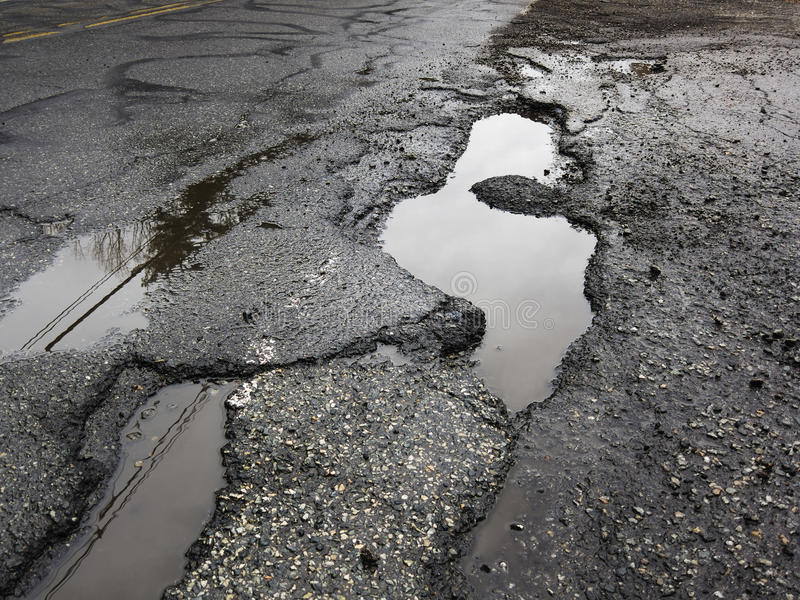 Une route avec au moins 3 nids de poule remplis d'eau.  La surface de la route est mouillée.  La route a l'air d'avoir de nombreuses plaques parce que la surface de la route a des textures différentes.