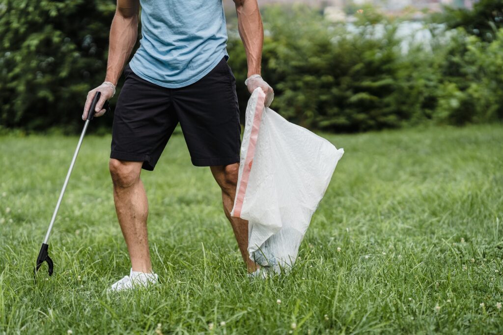 Photo of a person wearing black shorts and a blue t-shirt who is walking on grass. They are holding a white garbage bag and a tool to pick up litter. Photo d'une personne portant un short noir et un t-shirt bleu qui marche sur l'herbe. Elle tient un sac poubelle blanc et un outil pour ramasser les déchets.