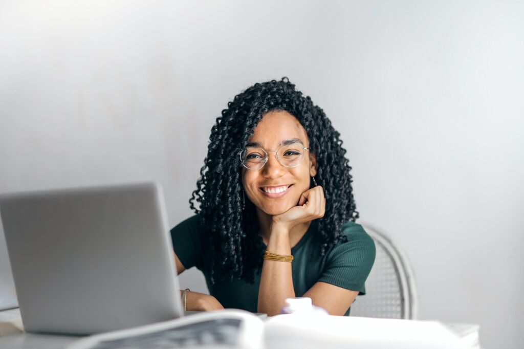 A woman sitting at a table with her laptop open.  She is sitting forward with her chin on her hand, smiling directly at the camera.  The woman has shoulder-length black curly hair and is wearing a green shirt and glasses.