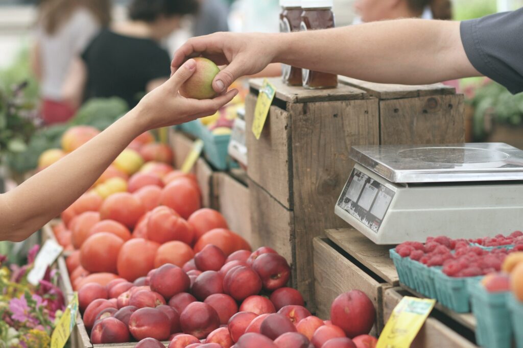 Photograph of a person giving an apple to a worker at a farmer's market.  Their arms reach across a large display of fresh fruit and vegetables including nectarines, tomatoes and raspberries.