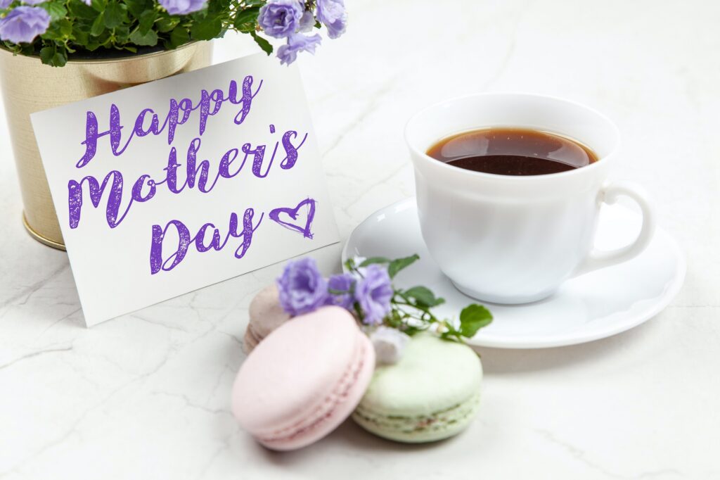 A photo of a card that says "Happy Mother's Day" sitting in front of a gold flower pot with a plan with lilac coloured flowers.  In front of the card is a teacup filled with coffee sitting on a saucer.  Both are white.  Beside the cup and saucer are 2 pink and 1 green macaron cookies.