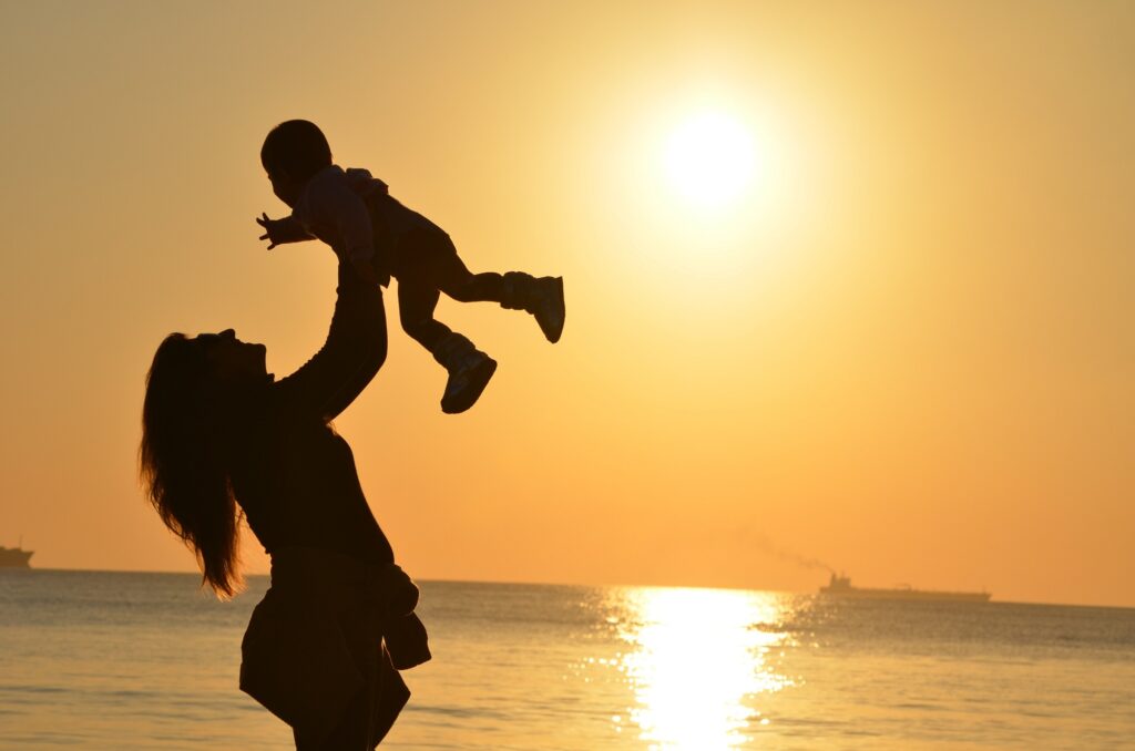 a silhouette photograph of a woman holding a toddler up in the air with a  yellow/orange sunset in the background.  It appears they are standing on a beach in front of a large body of water.