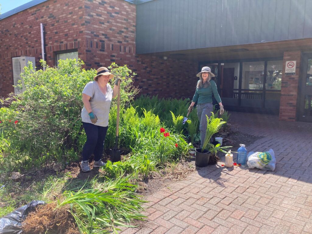 A photo of two community members working on the pollinator garden in front of Orleans Wood Elementary School.  They are wearing hats and gardening gloves.  They have shovels  in their hands and there are pots of plants around them.
