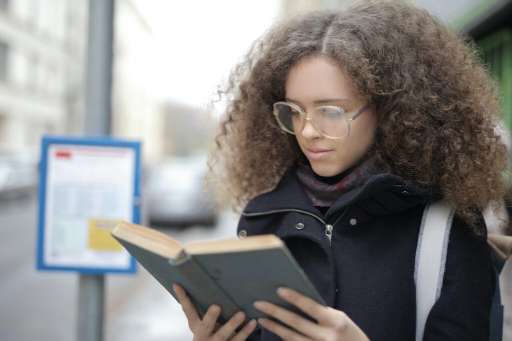 woman standing outside holding an open book.  She has long curly brown hair and is wearing glasses, a coat and has a strap from a bag slung on her shoulder.