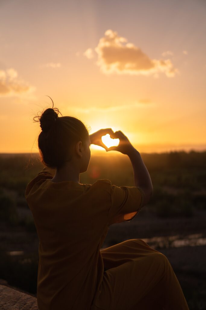 young woman sitting in front of a sunset.  She has her hands in the shape of a heart in front of the sun.