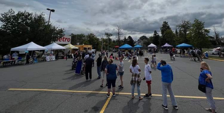 Photograph of the community barbeque site.  There is a long line up of people in the centre who are waiting to get food at Golden Fries.  Along the left side of the photo, there are tents lined up in a row leading to the food truck.  Along the right, towards the back of the site, there are more tents with activities and entertainment for attendees.