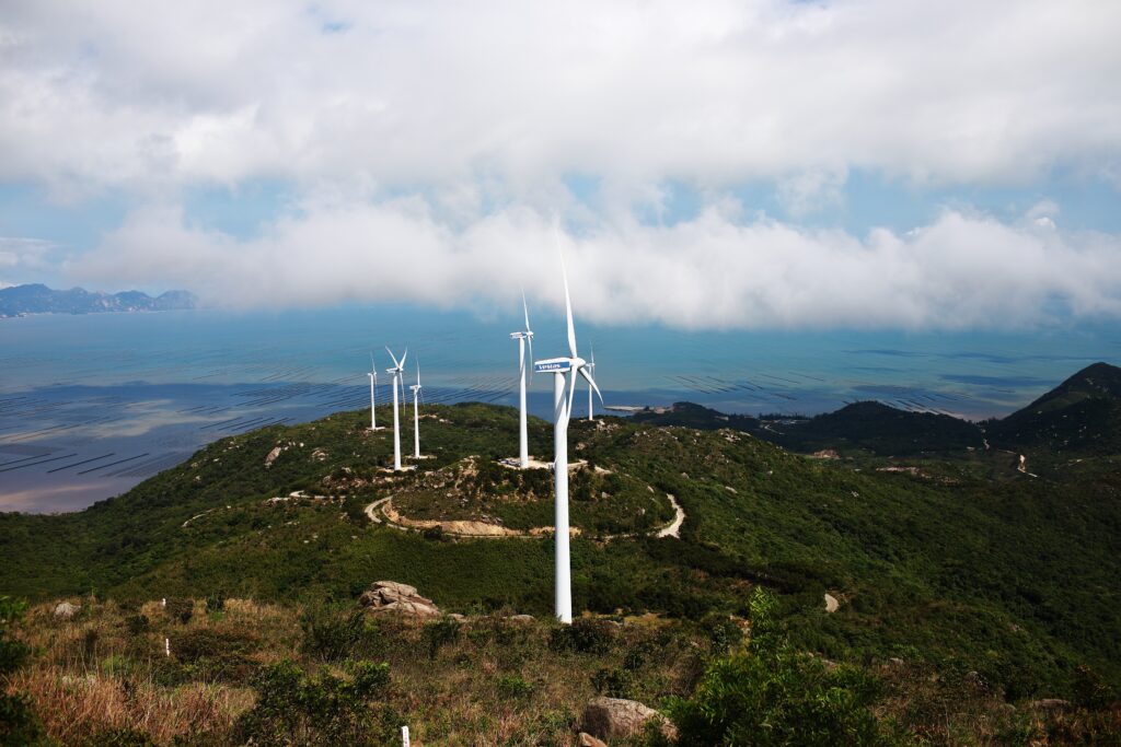 Photograph of large white windmills on the land along a coastline.  The land around the windmills is green and lush.  You can see clear blue water toward the back of the photograph.  There are fluffy white clouds in the sky.