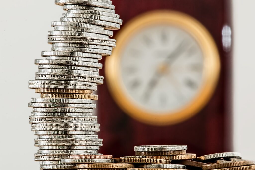 a photo of about 30 coins stacked vertically so you can see the edges of the coins with their ridges.  The coins are silver in colour but a few look tarnished and have a more copper colour.  There is a blurry background showing a clock behind the stack of coins.