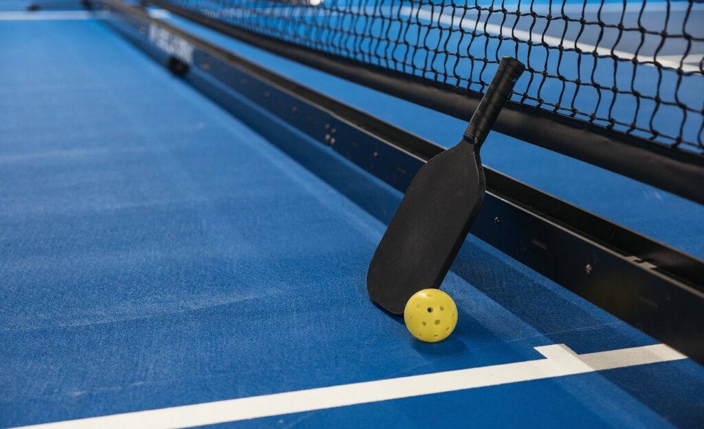 A photo of a black pickleball paddle resting against a black net. There is a yellow pickleball sitting beside the racket. The court surface is bright blue with white lines.