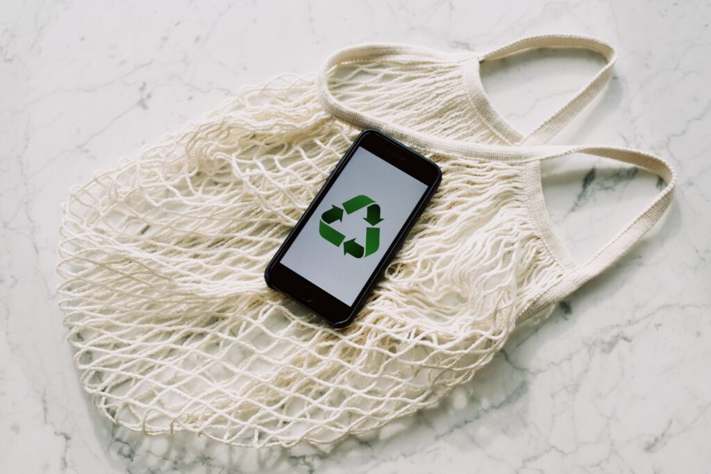 A photo of an undyed mesh shopping bag laying on a marble counter top. Sitting on top of the bag is a black smartphone. The smartphone is displaying the reduce, reuse, recycle icon (three green arrows in the shape of a triangle.