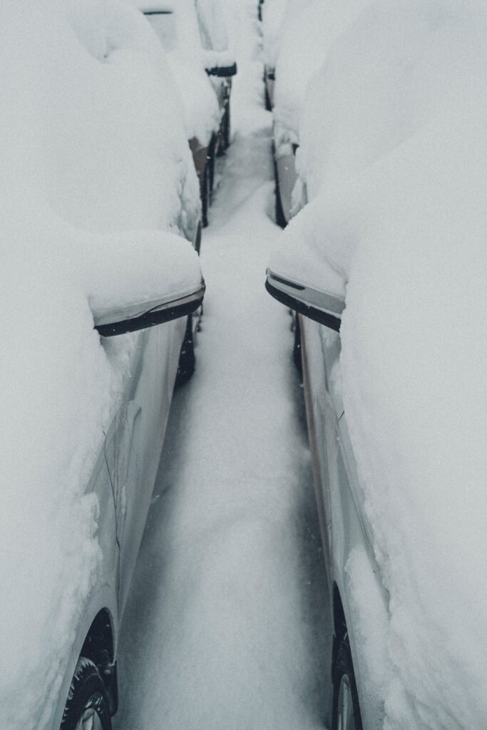 A photo of two white cars parked side by side in a parking lot. The cars are covered with snow. There is also a layer of snow on the ground.