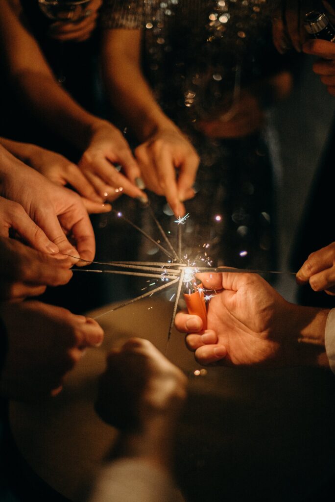 A photograph of a group of people - 9 hands reaching towards the centre with a sparkler. In the centre is a lighter lighting the sparklers.