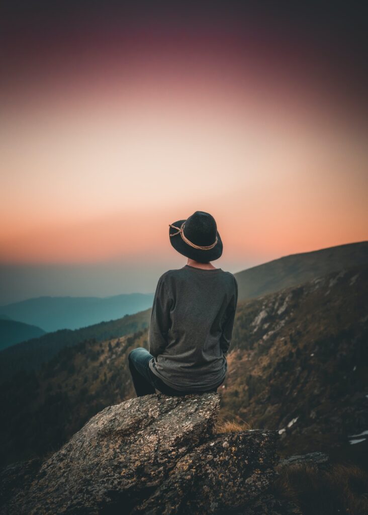 A photograph of a person from the back who is sitting on a rocky outcrop. They are looking out over the mountains, toward the peachy-coloured sky.
