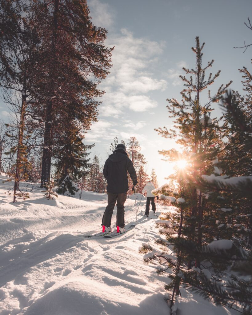A photograph of two people skiing on a forest trail. The sun is low in the sky. There is fresh snow on the trail.