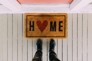 A photograph of someone looking down at their feet standing in front of a door mat at an entrance to a house. The door mat says "HOME" and the "O" is a heart.