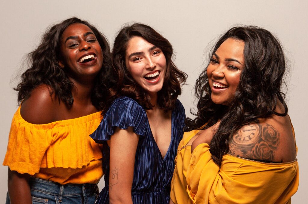 Three women standing together with wide smiles.  Two women are wearing bright golden yellow tops. The middle woman has a dark blue top.  