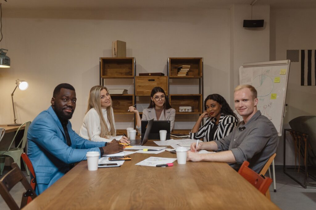 A group of 5 people sitting around a board room table. They have papers and pens on the table. There is a flip chart in the back that they've been drawing on.