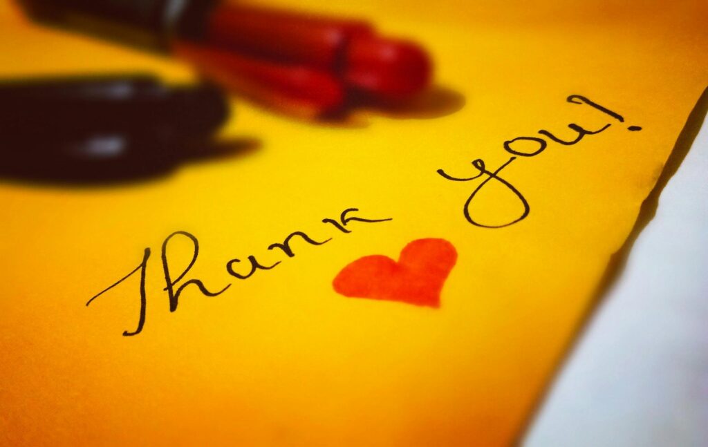 a photograph of a yellow piece of paper with black script saying Thank You! with a red heart drawn underneath.  In the background you can see the red and the black pens that were used.