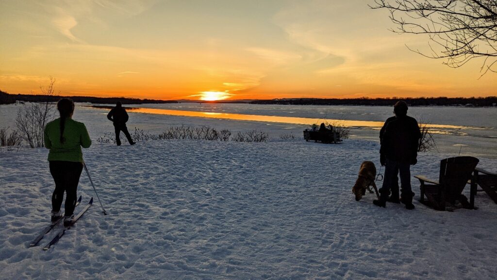 A photograph of the lookout point along the Ottawa river pathway near Green's Creek. There are three people in the photo. One standing at the cliff edge, looking toward the orange sunset over the Ottawa River. A second person is on skiis and standing further back. A third person is standing near the Adirondak chairs. There is snow on the ground.