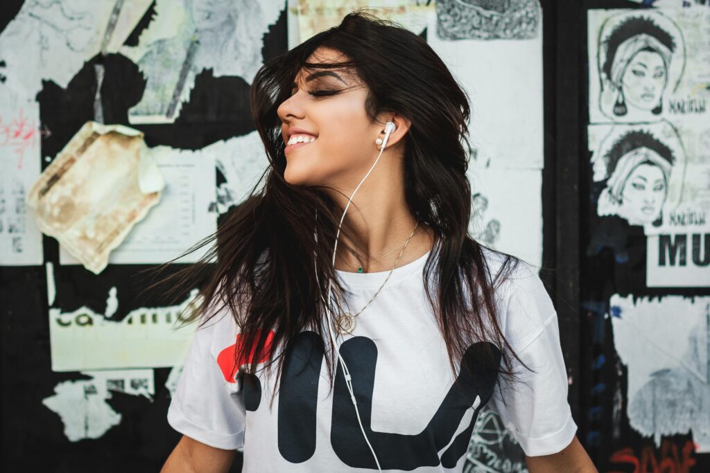 Young woman with long dark hair who is listening to music through earbuds. She has her eyes closed and is smiling. She's leaning against a wall that is covered in posters in various states of decay. She's wearing a white t-shirt with black and orange graphics on it.
