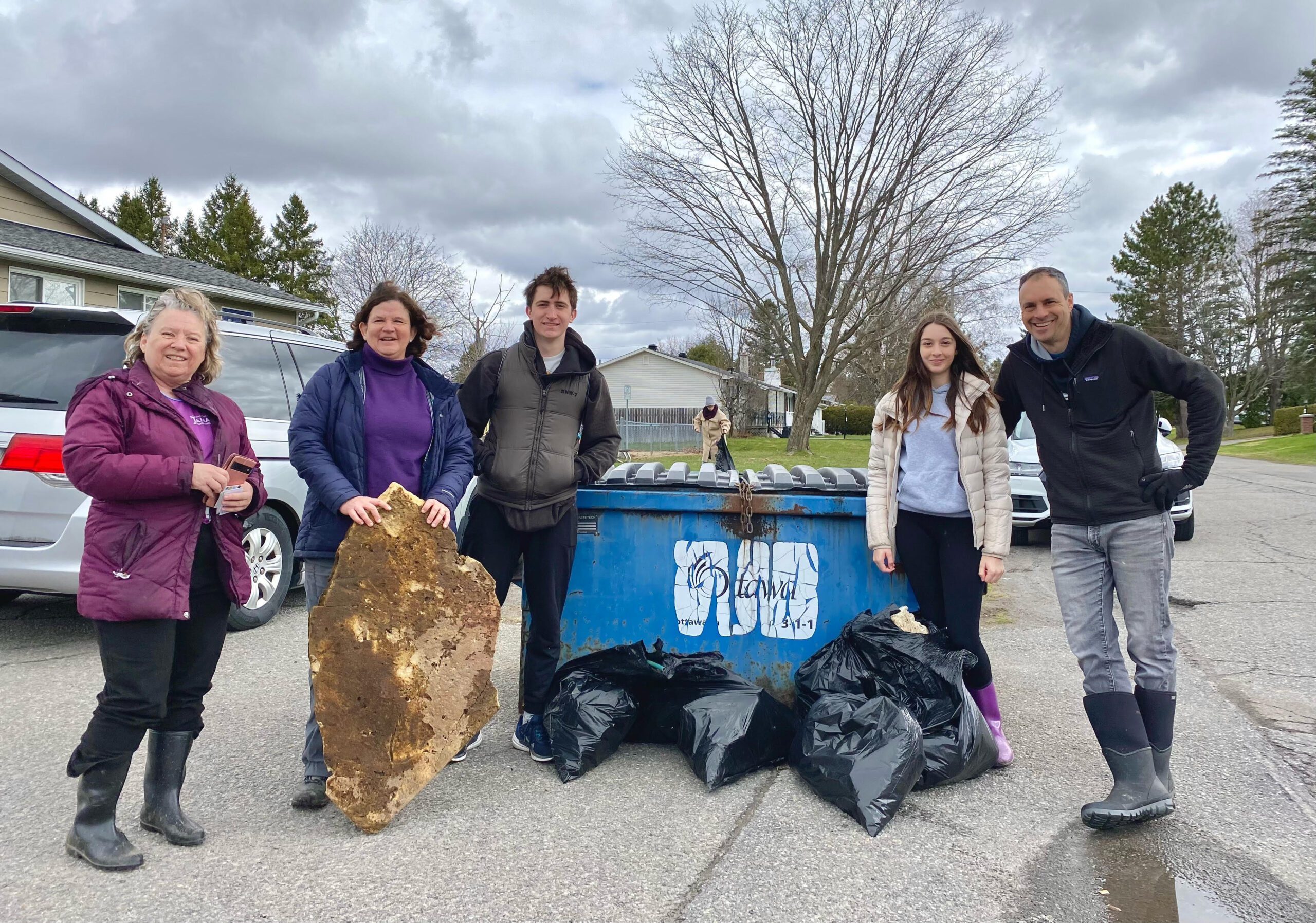 A photo from the 2024 Spring Clean Up - 5 community members stand around a blue dumpster and about 6 bags of trash that were collected at the clean up.